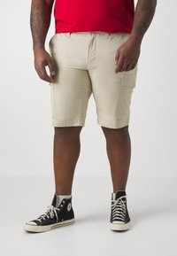 Pantaloni cargo beige con una vestibilità rilassata, dotati di tasche laterali e dettagli cuciti, abbinati a sneaker alte nere.