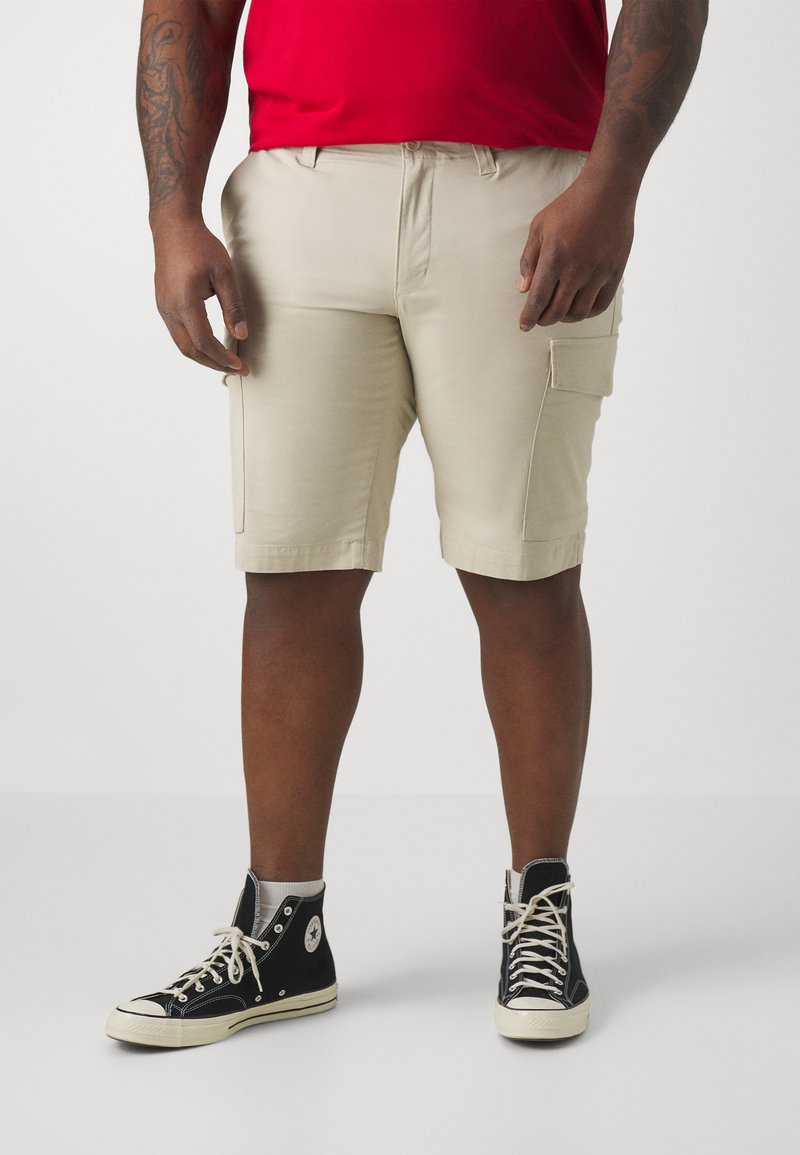Pantaloni cargo beige con una vestibilità rilassata, dotati di tasche laterali e dettagli cuciti, abbinati a sneaker alte nere.