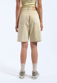 Bermudas de mezclilla en beige claro, con talle alto, diseño de cinco bolsillos y una etiqueta de marca de cuero en la parte trasera. Tejido texturizado.