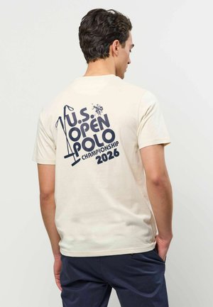 Mężczyzna o ciemnych włosach, ubrany w kremowy t-shirt z granatowym napisem "U.S. Open Polo Championship 2026" i grafiką kija polo na plecach.