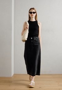 Black sleeveless top paired with a long black denim skirt and white platform sandals, holding a cream-colored bag. Smooth textures and minimalistic design.