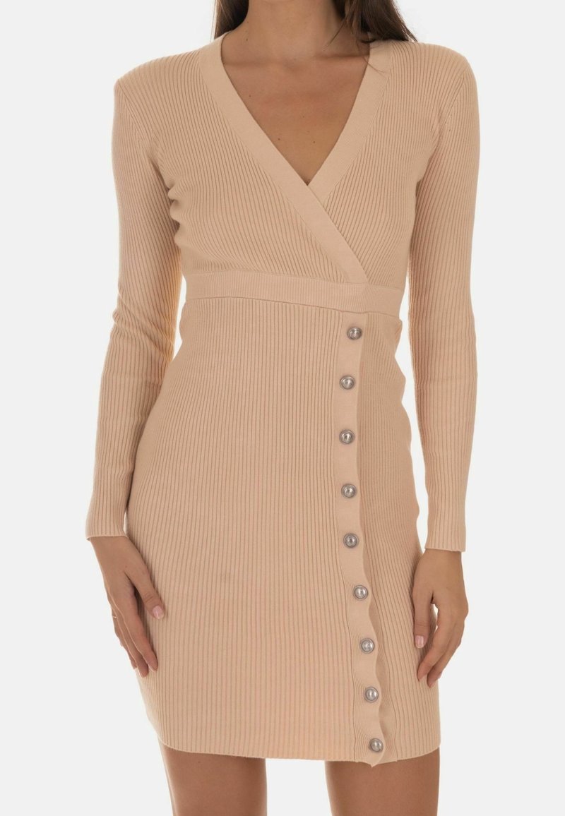 Femme portant une robe en maille côtelée beige à manches longues, col en V et une rangée de boutons sur le côté droit devant.