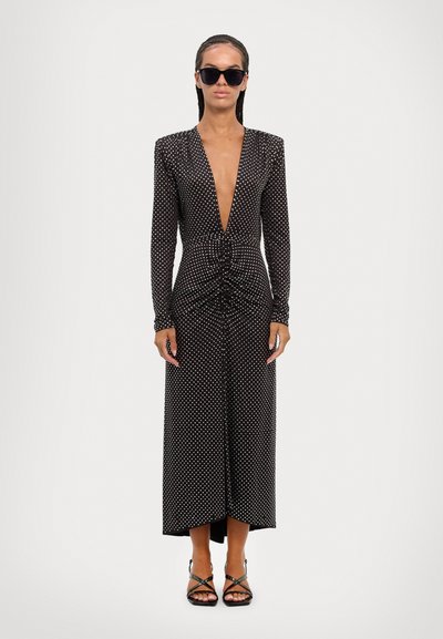 Lang zwarte jurk met witte stippen, diepe V-hals, lange mouwen, gathering in de taille en asymmetrische zoom. Gecombineerd met zwarte zonnebril en sandaaltjes met bandjes.
