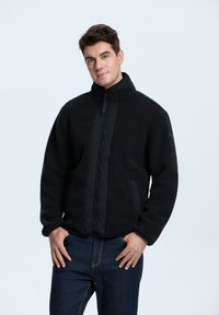 Mustat fleece-takki, jossa on korkea kaulus, vetoketjullinen etuosa ja sivutaskut. Ominaisuuksiin kuuluu teksturoidut pinnat ja sileät kangastuotteet.