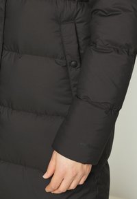 Crna puffer jakna s kabanom dizajnom, koja ima šaltersku džepu i vidljivi Patagonia logo na manžetama. Meki, topli materijal.