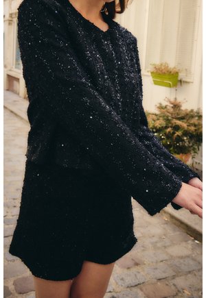 Vrouw draagt een zwart, lang mouwen top met pailletten en bijpassende korte broek, staand op een kasseienstraat met planten op de achtergrond.