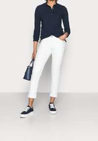 Marinblå långärmad polo skjorta, vita jeans med rullade ärmar, marinblå sneakers med vita snören och en liten marinblå handväska. Platt, neutral bakgrund.