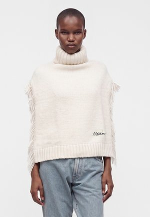 Personne portant un poncho en tricot crème à col roulé avec des franges sur les côtés et un jean bleu clair sur un fond uni.