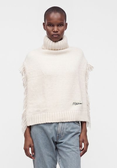 Personne portant un poncho en tricot crème à col roulé avec des franges sur les côtés et un jean bleu clair sur un fond uni.