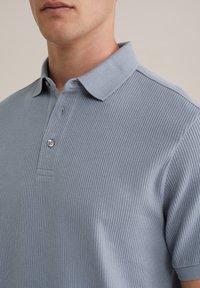 Cămașă polo cu un voal albastru deschis, dotată cu un guler clasic, plachetă cu trei butoane și o textură subtilă pe întreaga țesătură.