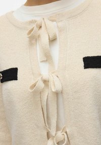 Beige gebreide cardigan met een zachte textuur, voorzien van een sluiting met een strik aan de voorkant en zwarte geribde accenten op de zakken.