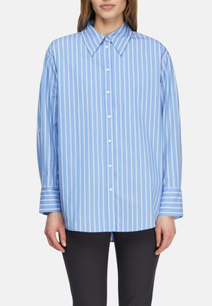 Kobieta w niebieskiej koszuli na guziki z białymi pionowymi prążkami i dużymi mankietami, ubrana w czarne spodnie, stojąca na białym tle.