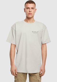 Lys grå oversized t-shirt i bomuld med korte ærmer, med arabisk tekst og den engelske sætning "Elsk dig selv først" trykt på forsiden.