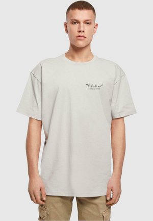 Lys grå, oversized t-skjorte i bomull med korte ermer, med arabisk tekst og engelsk frase "Elsk deg selv først" trykt på forsiden.