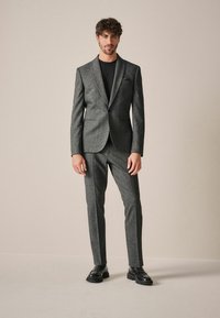 Traje gris con un diseño ajustado, que incluye un blazer de un solo botón y pantalones a medida a juego. Zapatos negros completan el look.