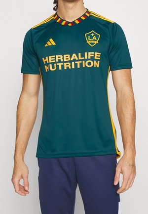 Man wearing dark teal LA Galaxy soccer jersey with yellow accents and "HERBALIFE NUTRITION" sponsor logo, paired with navy athletic shorts.