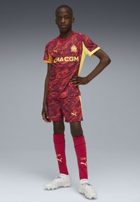 Maglia da calcio a righe rosse e gialle con pantaloni abbinati. Calze rosse fino al ginocchio. Scarpetta da calcio bianche con superficie testurizzata e lacci.