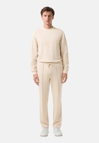 Beiger Sweatshirt und passende Jogginghose aus einem weichen Stoff; verfügt über klare Linien, eine lockere Passform und weiße Sneaker.
