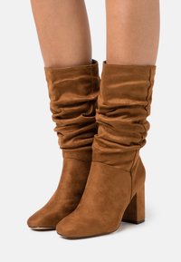 Stivali alla caviglia marroni, morbidi, realizzati in pelle scamosciata, con punta affusolata e tacco spesso per una maggiore stabilità. Il tessuto texturizzato aggiunge interesse visivo.