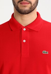 Rotes Polohemd aus strukturiertem Stoff, mit klassischem Kragen und drei weißen Knöpfen, verziert mit einem grünen Krokodil-Logo auf der Brust.