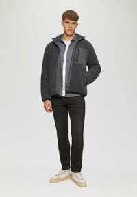 Grijze fleece jas met capuchon, gestructureerde stof en een borstzak. Draagt over een denim shirt, gecombineerd met donkere jeans en lichte sneakers.