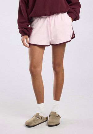 Personne portant un short à cordon de serrage rose pâle avec des bordures bordeaux, des chaussettes blanches et des chaussures beige à enfiler, debout devant un fond uni.