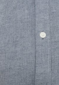 Primer plano de una tela de mezclilla azul clara con una costura vertical y un solo botón blanco de cuatro agujeros cosido cerca del centro.