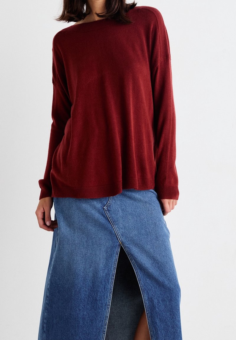 Femme portant un pull ample rouge foncé et une jupe en denim bleue avec une fente devant, sur un fond uni clair.