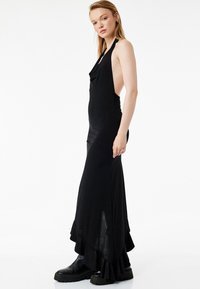 Schwarzes Maxikleid mit Neckholder, gerafftem Vorderteil, offenen Seiten und gerafftem Saum. Kombiniert mit klobigen schwarzen Stiefeln, glatte Textur.