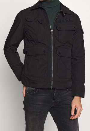Homme portant une veste noire zippée avec plusieurs poches boutonnées sur un col roulé vert foncé et un jean sombre, debout avec une main dans la poche.