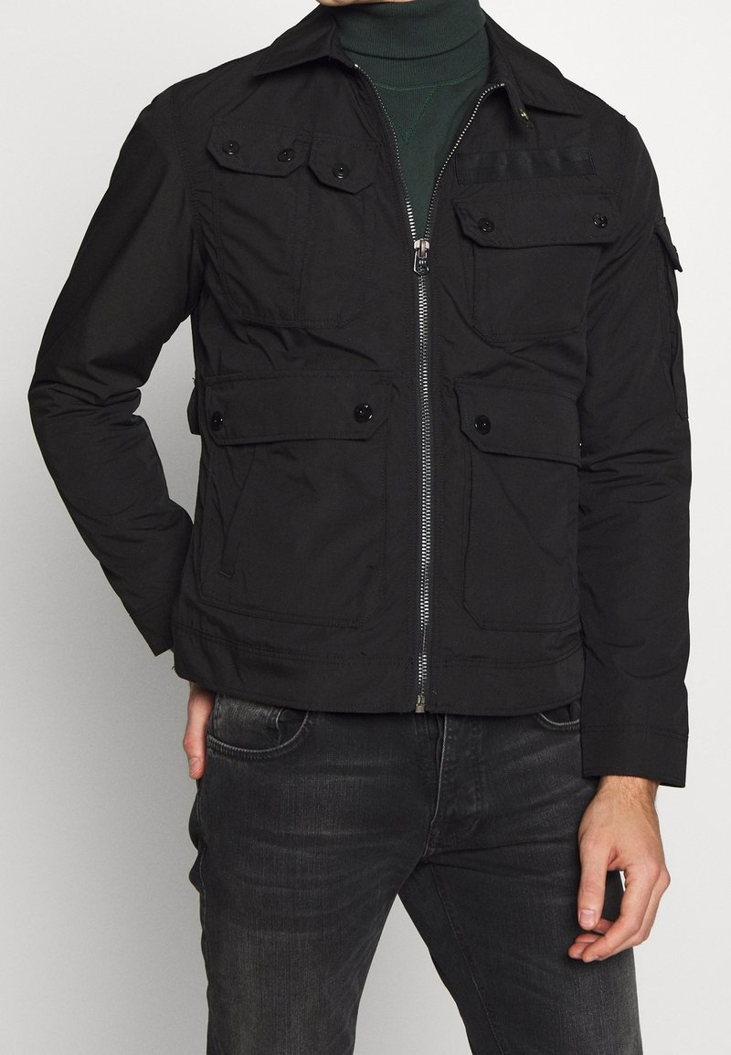 Homme portant une veste noire zippée avec plusieurs poches boutonnées sur un col roulé vert foncé et un jean sombre, debout avec une main dans la poche.