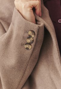 Der Ärmel eines beigen Wollmantels mit weicher Textur verfügt über drei goldene Knöpfe mit dekorativen Löchern und eine ordentlich umgeschlagene Manschette.