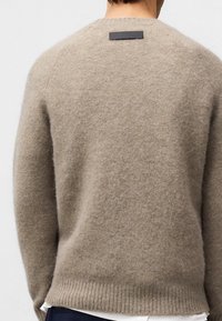 Beżowy sweter wełniany z teksturowanym wykończeniem, żebrowanym dołem i małą prostokątną czarną skórzaną metką na plecach przy dekolcie.