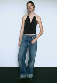 Czarny top na ramiączkach z drapowanym dekoltem i srebrnymi wstawkami, zestawiony z niebieskimi szerokimi spodniami dżinsowymi z podwiniętymi nogawkami i klasycznym pięciopokoleniowym krojem.