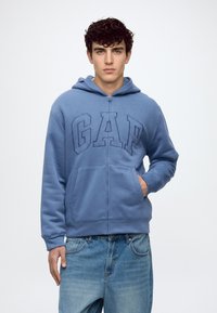 Jeune homme aux cheveux bouclés foncés, portant un sweat à capuche zippé bleu GAP et un jean bleu, debout avec une main dans la poche.