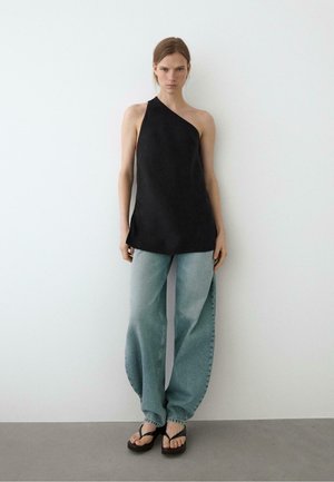 Frau steht vor weißer Wand, trägt schwarzen Ein-Schulter-Top, lockere hellblaue Jeans und schwarze Plateausandalen, schaut nach vorne mit neutralem Gesichtsausdruck.