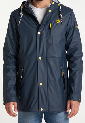 Mężczyzna ubrany w granatową, wodoodporną kurtkę z żółtymi guzikami, suwakami i sznurkami kaptura, z małym żółtym logo na piersi.