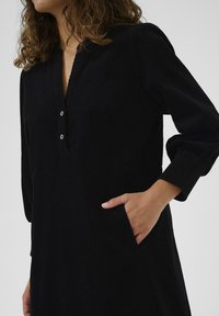 Black dress with a V-neck and three-quarter sleeves, featuring a placket with two buttons and side pockets, made of textured fabric.