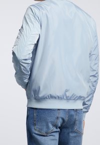 Jasnoniebieska nylonowa kurtka bomberka z ściągaczami przy mankietach i na dole, z delikatną teksturą oraz zapinaną na zamek boczną kieszenią na lewym rękawie.