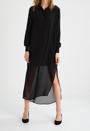 Kobieta w długiej czarnej przezroczystej sukni z długimi rękawami i wysokimi rozcięciami po bokach, w czarnych szpilkach, na białym tle.