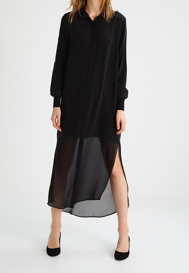 Femme portant une longue robe noire transparente à manches longues et fentes hautes sur les côtés, associée à des talons hauts noirs sur fond blanc.