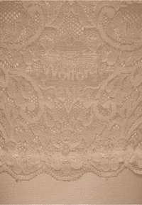 Tessuto di pizzo beige con motivi floreali intricati e bordi scallop. Il nome del marchio "Wolford" è delicatamente impresso nel design.