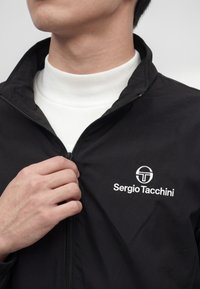 Person trägt eine schwarze Sergio Tacchini Jacke und öffnet teilweise den Reißverschluss über einem weißen, gerippten Rollkragenpullover.