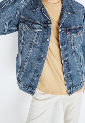 Persona che indossa una giacca di jeans blu sbottonata sopra una camicia beige e pantaloni bianchi, in piedi davanti a uno sfondo chiaro e uniforme.