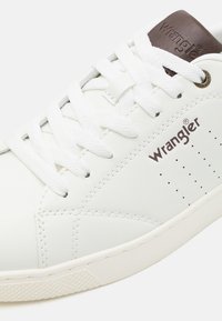 Baltas ādas treneris ar baltām auklām, piedāvājot brūnu akcentu uz papēža un perforētus detalizējumus sānos. Wrangler logotips brūnā krāsā.