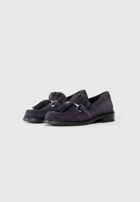Mocassins en daim gris foncé avec des accents de glands et des détails en métal argenté. Bout arrondi et talon bas, avec une semelle cousue pour plus de texture.