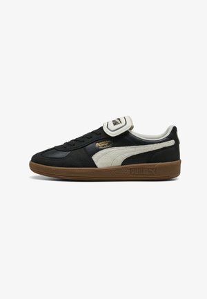Zapatilla Puma Palermo en blanco y negro con suela marrón, franja blanca con el logo de Puma, cinta en la lengüeta con la marca y texto dorado "Palermo" en el lateral.