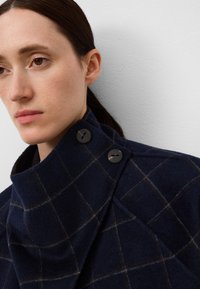 Cappotto di lana blu navy con motivo a scacchi, colletto alto e bottoni neri. Tessuto testurizzato con una finitura morbida. Primo piano dei dettagli del design.