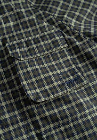 Tessuto a quadri verde e blu navy con una tasca a pattina cucita, caratterizzata da un piccolo logo ricamato nell'angolo inferiore.