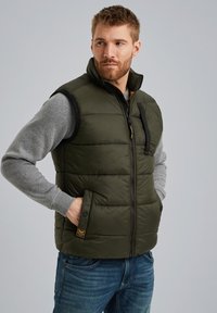 Olivgrüne Steppweste mit durchgehendem Reißverschluss, Seitentaschen und schwarzen Akzenten. Getragen über einem grauen Langarmshirt und Denim-Jeans.